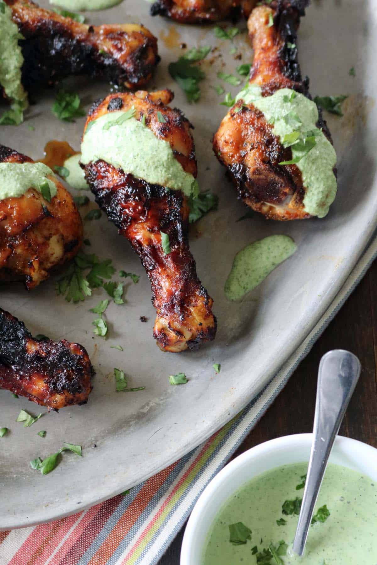 Air-Fried Peruvian Chicken with Green Crema drumsticks are arranged on a gray platter, topped with green herb sauce and chopped cilantro. A small bowl of the vibrant green crema with a spoon sits nearby.