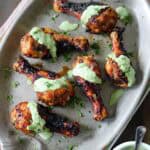 Air-Fried Peruvian Chicken with Green Crema drumsticks are arranged on a large oval platter, topped with vibrant green sauce and garnished with chopped herbs. A small bowl of extra green crema sits nearby on a striped cloth.