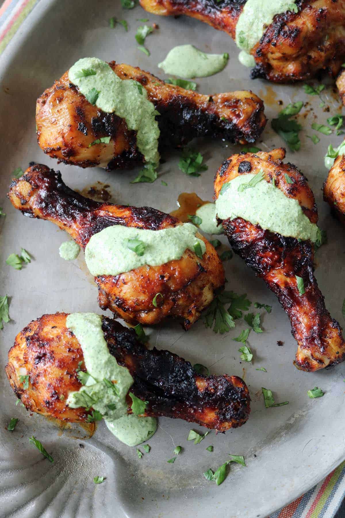 Grilled chicken drumsticks topped with a vibrant green herb sauce reminiscent of Air-Fried Peruvian Chicken with Green Crema, garnished with chopped fresh herbs and arranged on a gray serving platter.