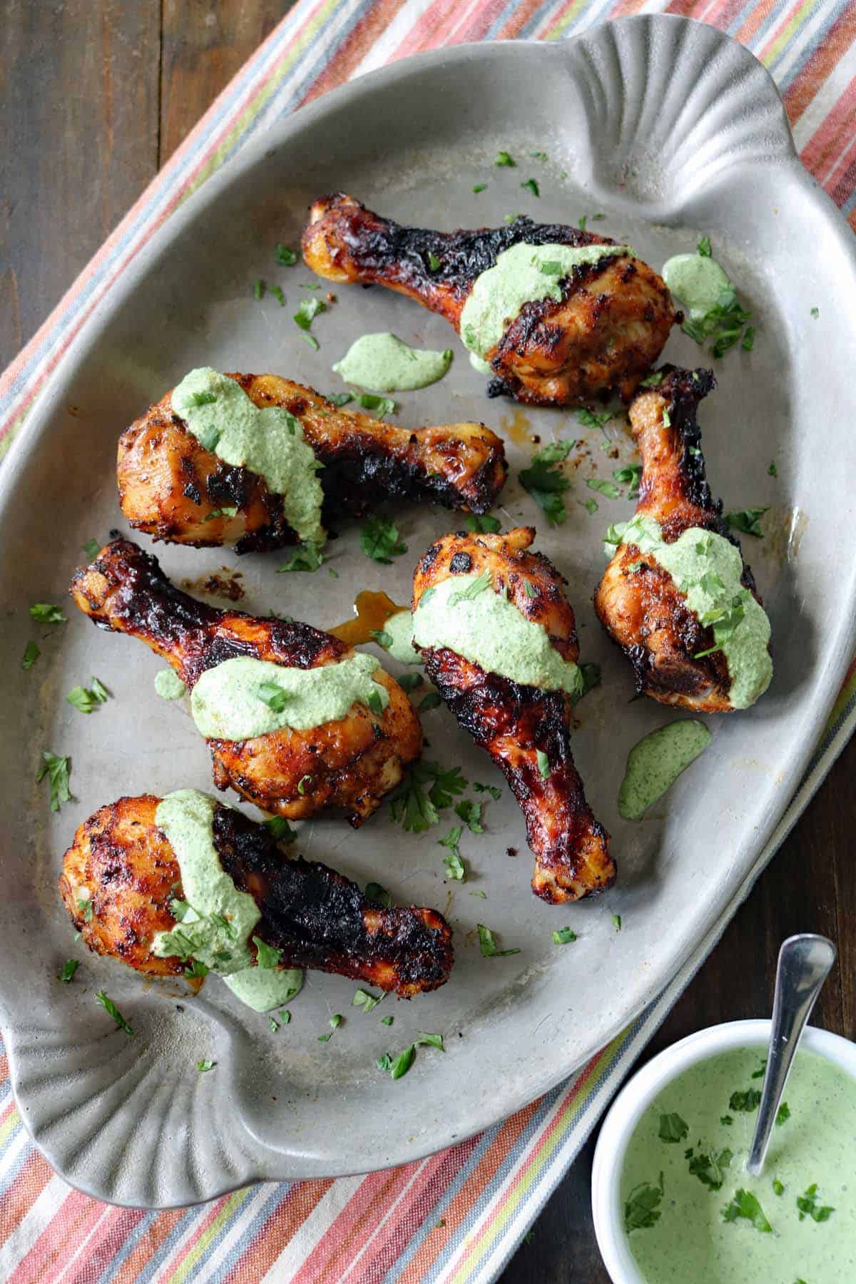 Air-Fried Peruvian Chicken with Green Crema drumsticks are arranged on a large oval platter, topped with vibrant green sauce and garnished with chopped herbs. A small bowl of extra green crema sits nearby on a striped cloth.