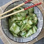 A bowl of Asian Cucumber Ribbon Salad with sesame seeds sits on a black-and-white patterned dish, placed on a bamboo mat. Red chopsticks with a floral design rest atop the vibrant cucumber salad.