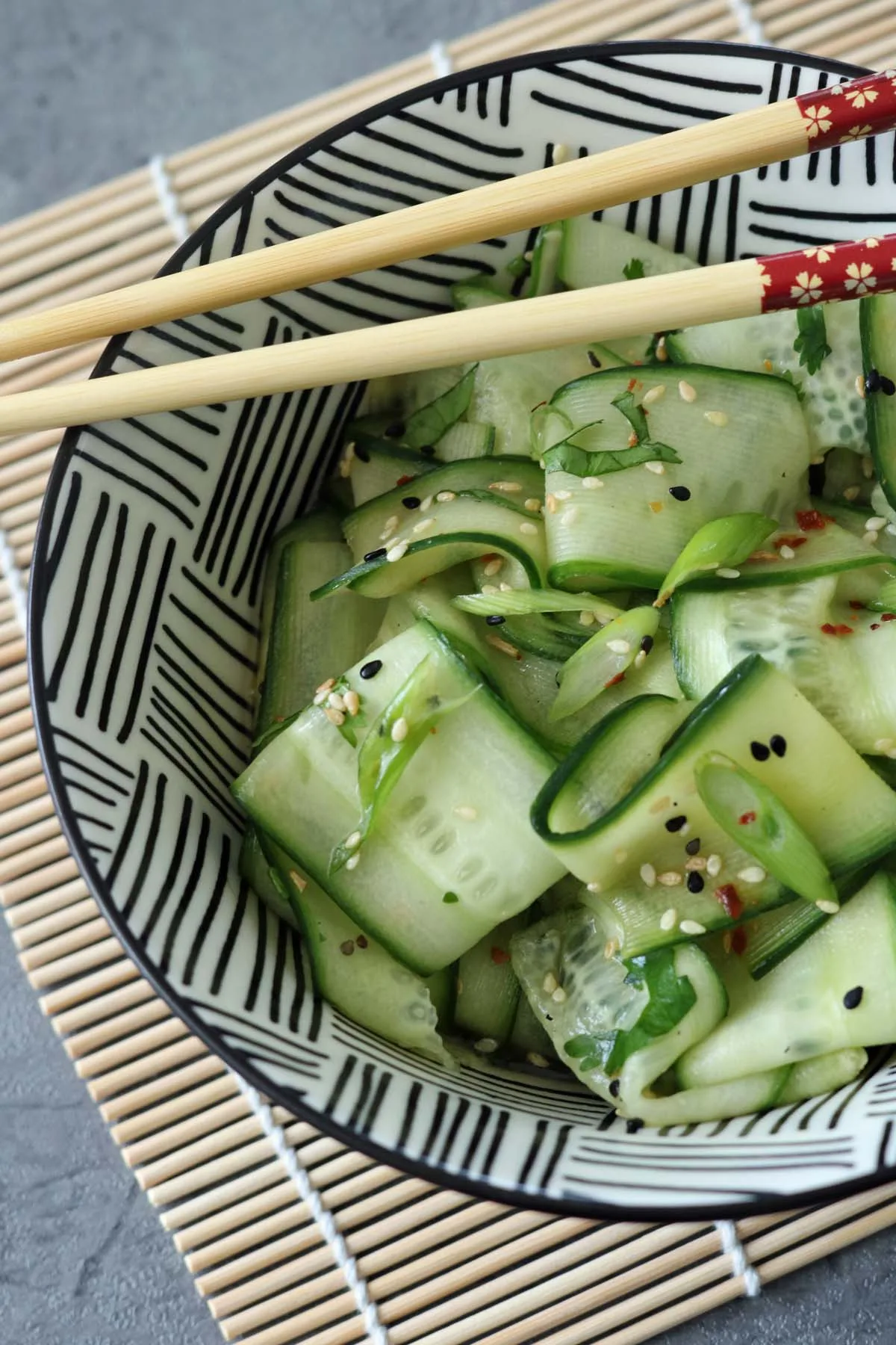 A bowl of Asian Cucumber Ribbon Salad with thinly sliced cucumber ribbons, sprinkled with sesame seeds, sits on a bamboo mat. A pair of chopsticks rests on the bowl’s rim, which features a black and white geometric pattern.