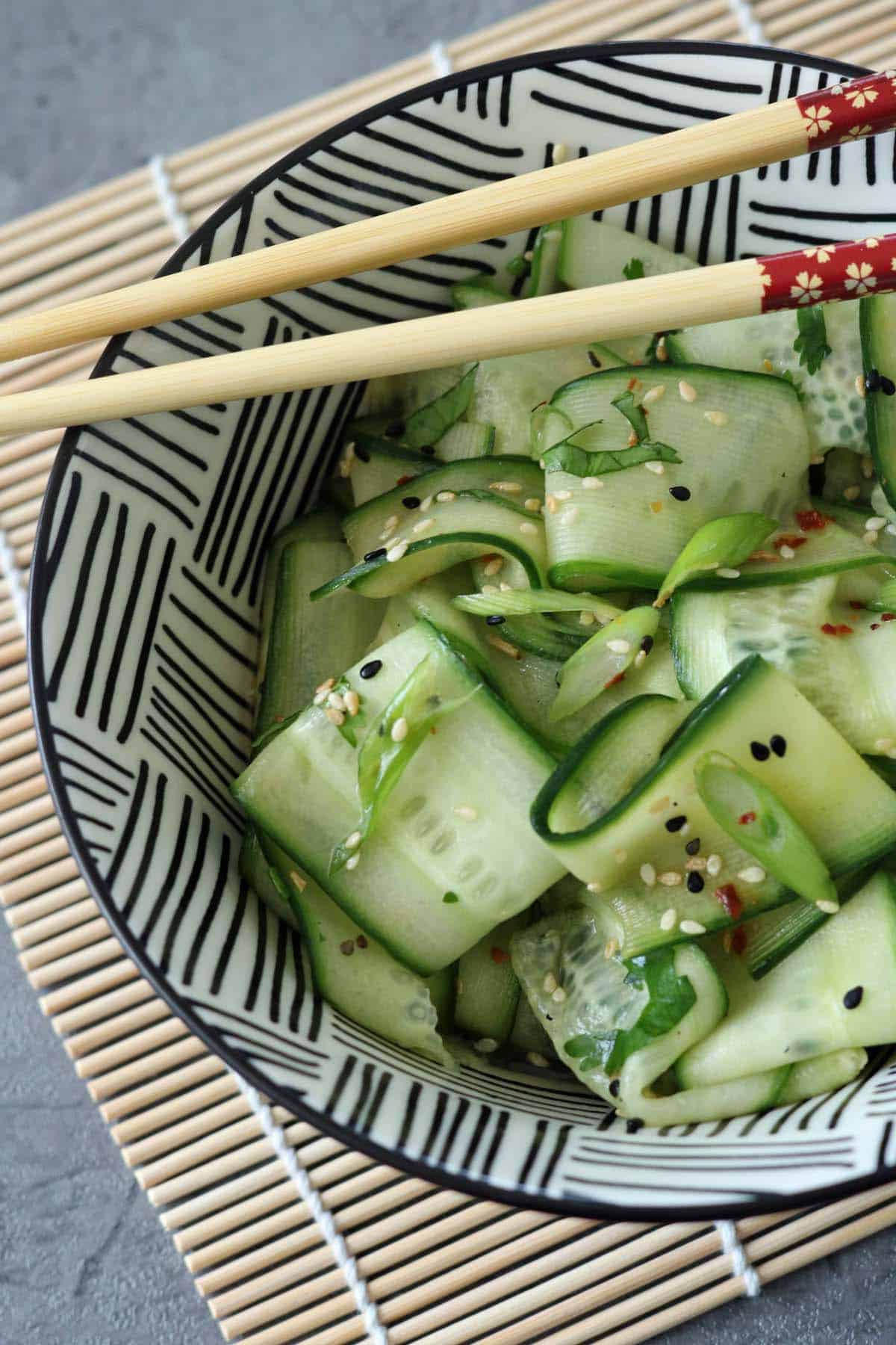 A bowl of Asian Cucumber Ribbon Salad with thinly sliced cucumber ribbons, sprinkled with sesame seeds, sits on a bamboo mat. A pair of chopsticks rests on the bowl’s rim, which features a black and white geometric pattern.