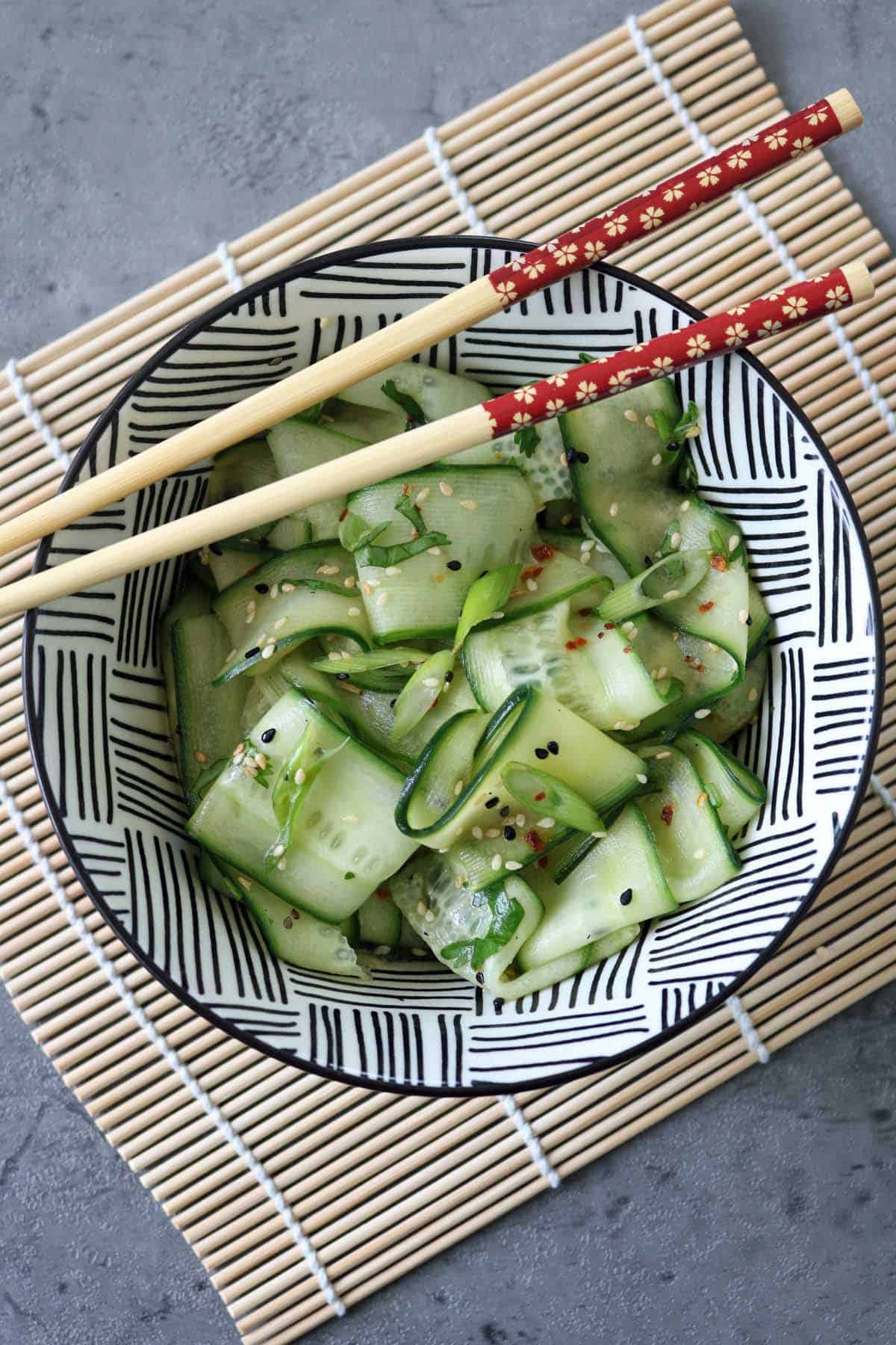 A bowl of Asian Cucumber Ribbon Salad with sesame seeds sits on a black-and-white patterned dish, placed on a bamboo mat. Red chopsticks with a floral design rest atop the vibrant cucumber salad.