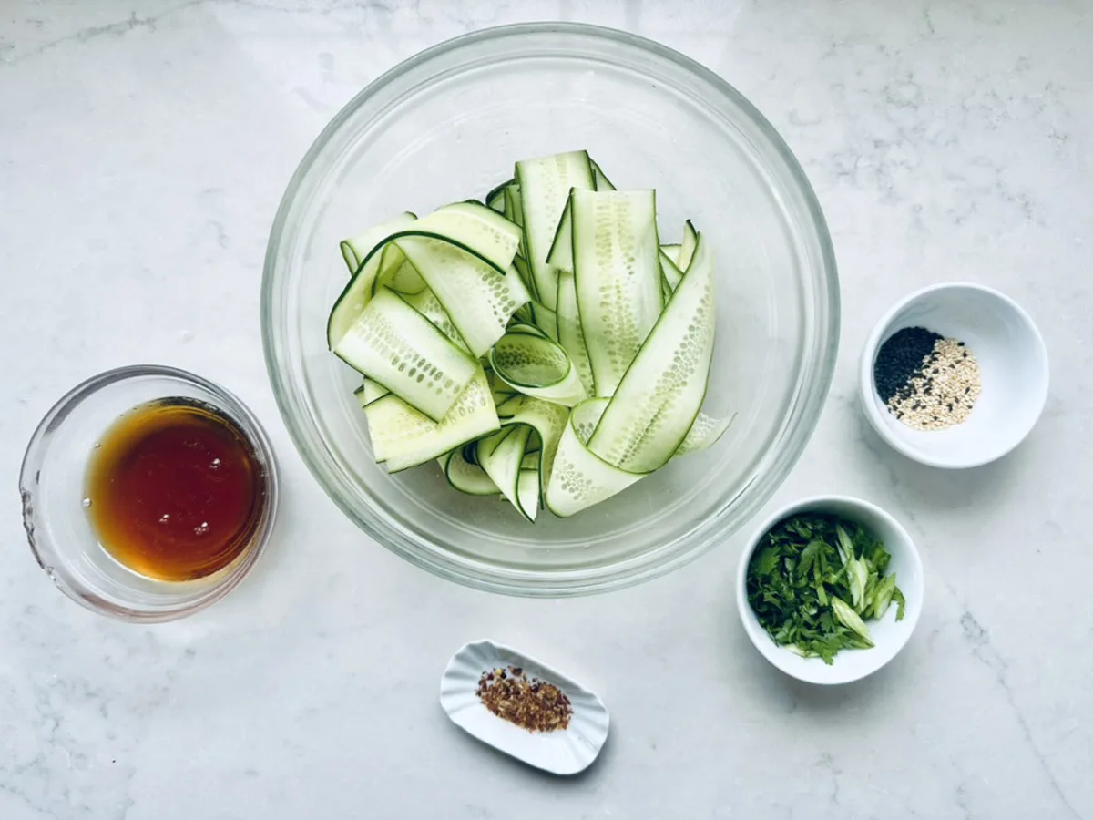 A glass bowl of Asian Cucumber Ribbon Salad sits on a white surface, surrounded by small bowls of sauce, chopped green onions, spices, and seasonings.
