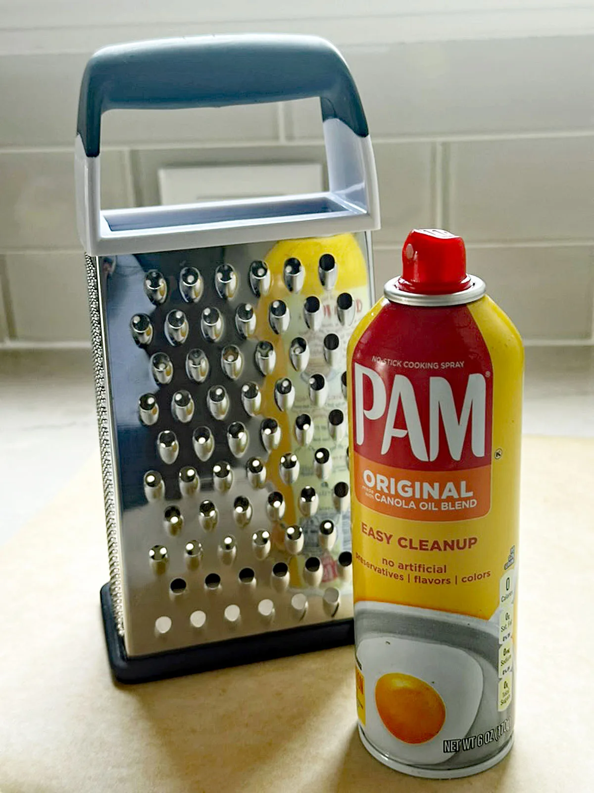 A metal box grater and a can of PAM Original cooking spray are placed on a kitchen counter with a tiled backsplash in the background.