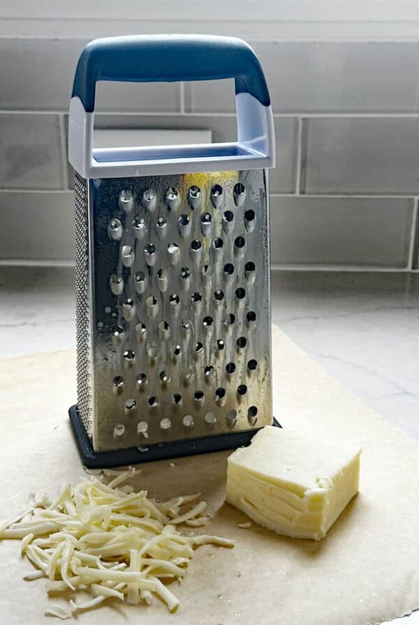 A box cheese grater stands on parchment paper next to a block of cheese and a small pile of grated cheese, with a tiled backsplash in the background.