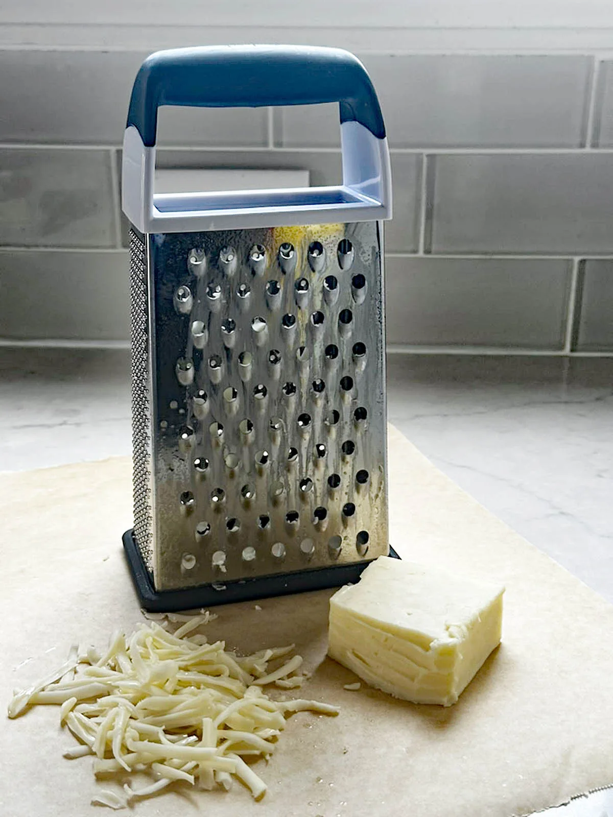 A box cheese grater stands on parchment paper next to a block of cheese and a small pile of grated cheese, with a tiled backsplash in the background.