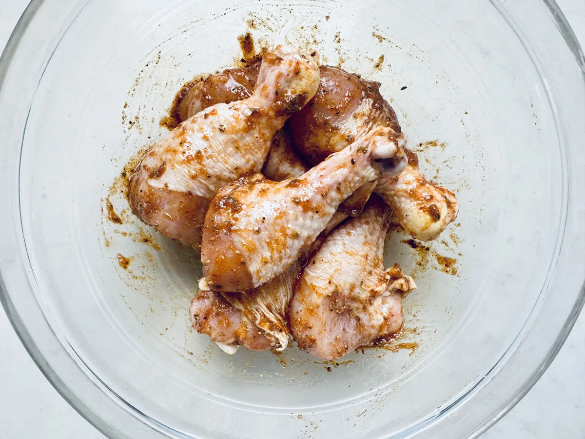 Raw chicken drumsticks coated with a reddish-brown spice marinade sit in a clear glass bowl, ready to become Air-Fried Peruvian Chicken with Green Crema. The bowl rests on a light-colored surface.