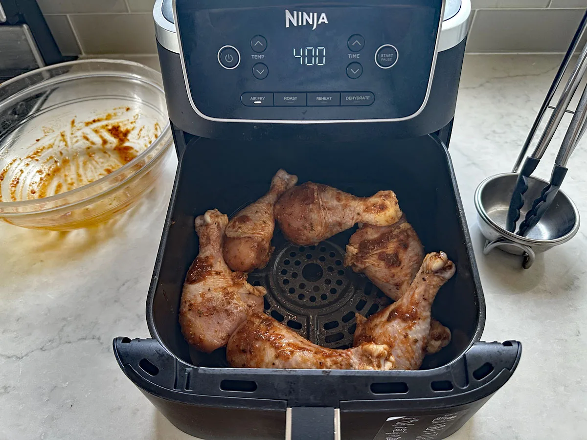 Five seasoned chicken drumsticks arranged in the basket of a Ninja air fryer set to 400 degrees, perfect for making Air-Fried Peruvian Chicken with Green Crema. An empty glass mixing bowl and metal tongs sit on the countertop nearby.