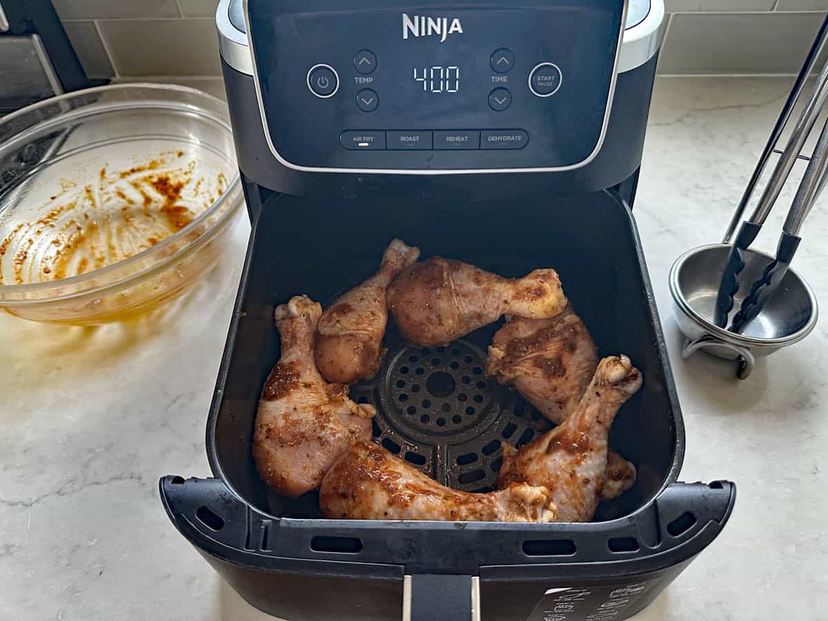 Five seasoned chicken drumsticks arranged in the basket of a Ninja air fryer set to 400 degrees, perfect for making Air-Fried Peruvian Chicken with Green Crema. An empty glass mixing bowl and metal tongs sit on the countertop nearby.