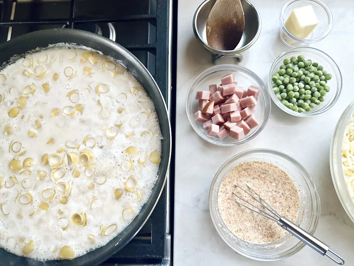 A pan of pasta cooking on a stove, next to bowls containing cubed ham, peas, butter, shredded cheese, cream, breadcrumbs, and a whisk on a light-colored countertop.