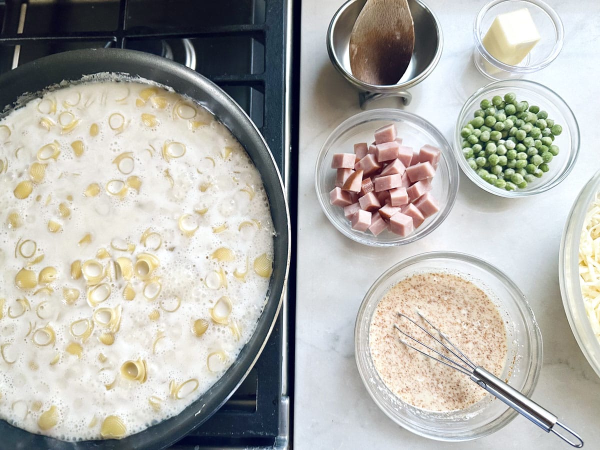 A pan of pasta cooking on a stove, next to bowls containing cubed ham, peas, butter, shredded cheese, cream, breadcrumbs, and a whisk on a light-colored countertop.