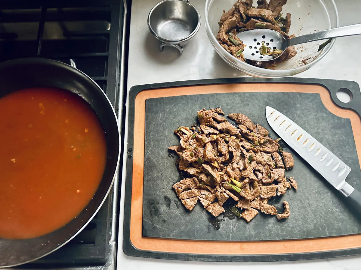 Sliced cooked beef on a cutting board with a chefs knife, a bowl of more beef pieces, a slotted spoon, and a pan of red sauce cooking on a stove. The scene suggests meal preparation.