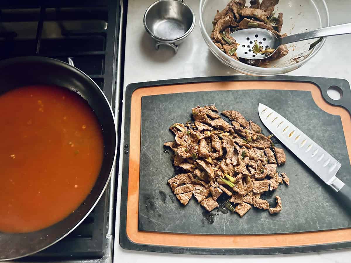 Sliced cooked beef on a cutting board with a chefs knife, a bowl of more beef pieces, a slotted spoon, and a pan of red sauce cooking on a stove. The scene suggests meal preparation.