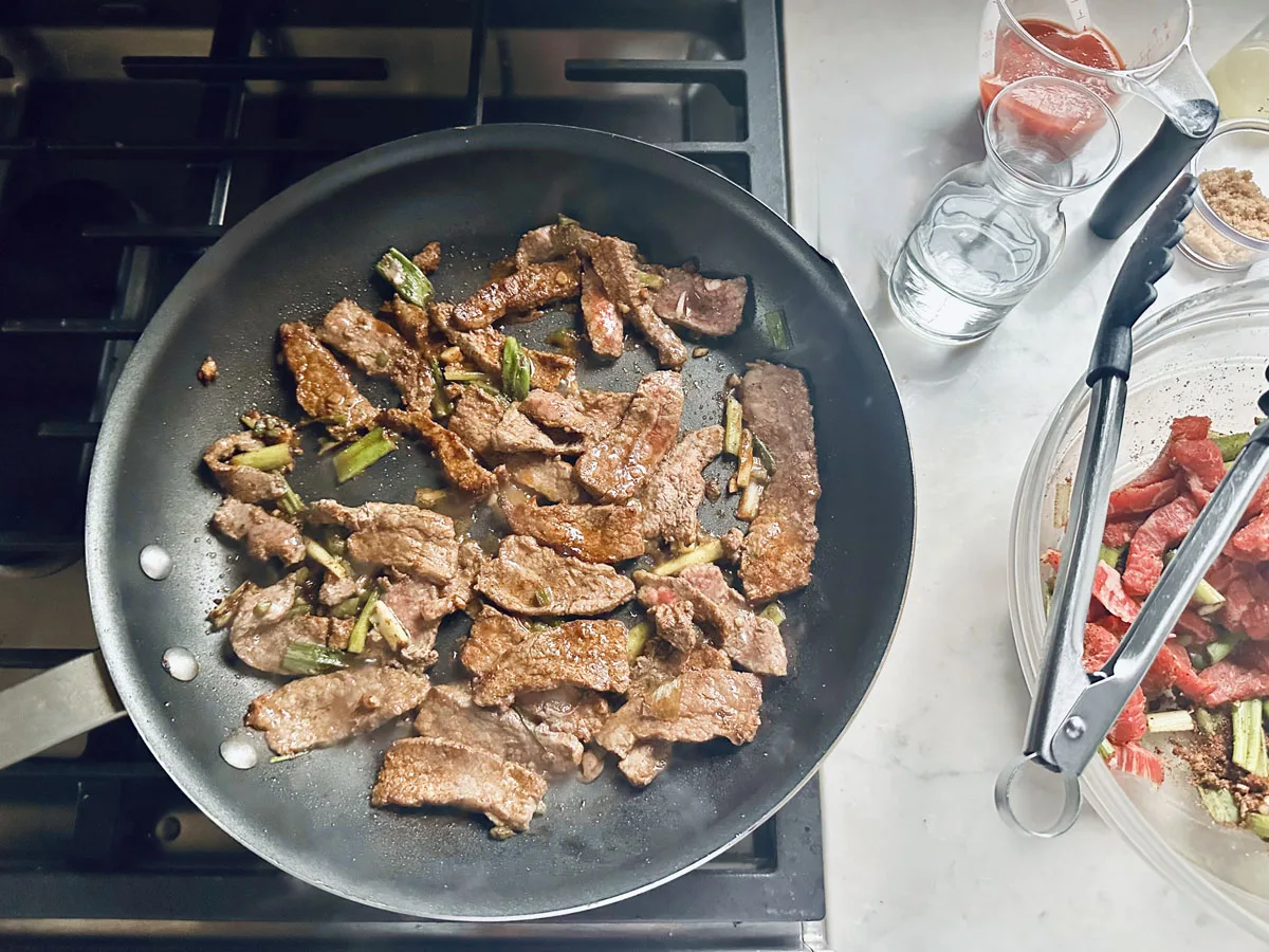 Strips of beef and green vegetables are cooking in a black skillet on a stovetop. Nearby, a glass bowl with uncooked beef strips, a whisk, and cups of sauce, water, and brown sugar are on the counter.