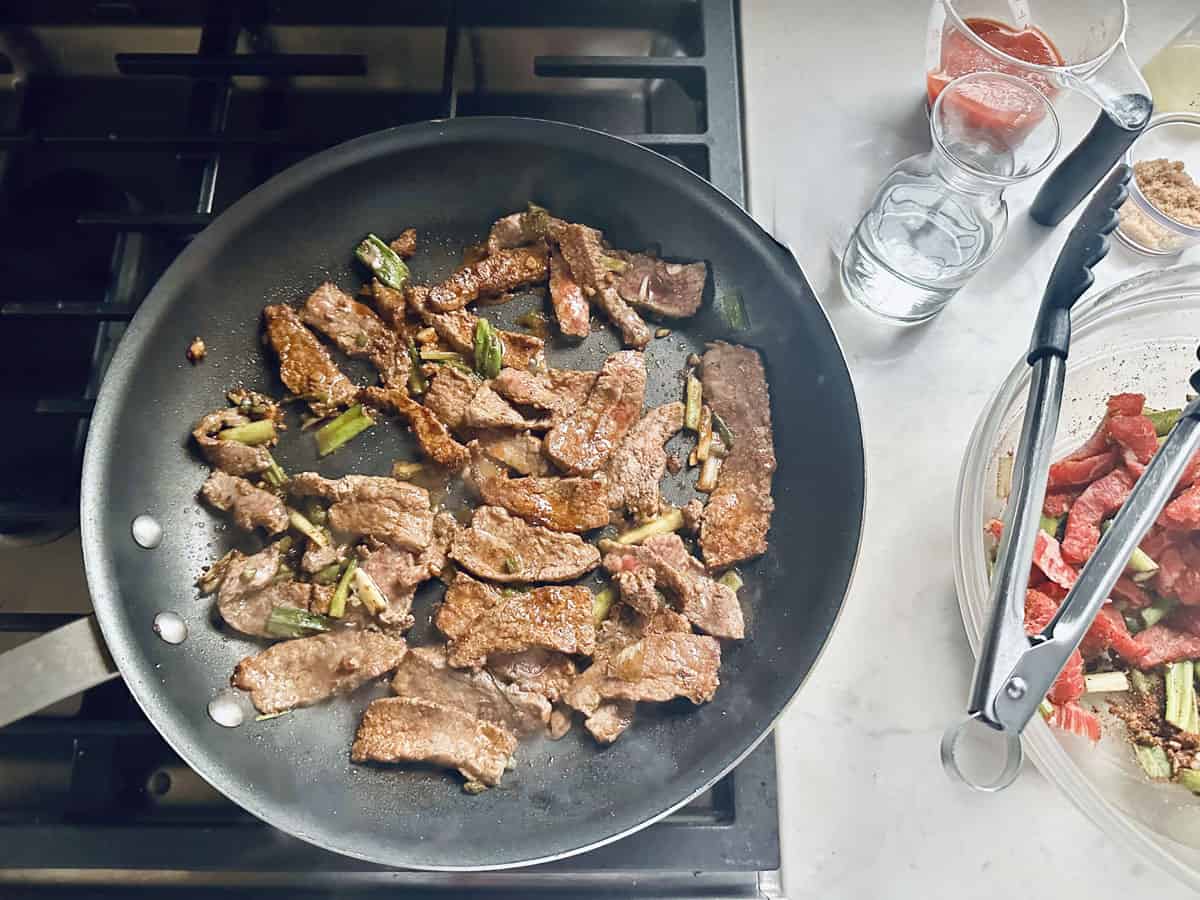 Strips of beef and green vegetables are cooking in a black skillet on a stovetop. Nearby, a glass bowl with uncooked beef strips, a whisk, and cups of sauce, water, and brown sugar are on the counter.