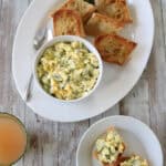 A white platter holds toasted bread slices and a bowl of Cucumber and Basil Egg Salad with herbs. A spoon rests on the side, and a green leafy garnish is placed on the platter. A small plate and a drink are beside it on a wooden table.