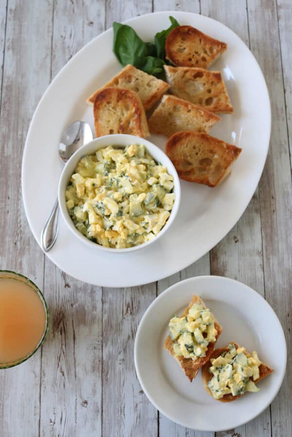 A white platter holds toasted bread slices and a bowl of Cucumber and Basil Egg Salad with herbs. A spoon rests on the side, and a green leafy garnish is placed on the platter. A small plate and a drink are beside it on a wooden table.
