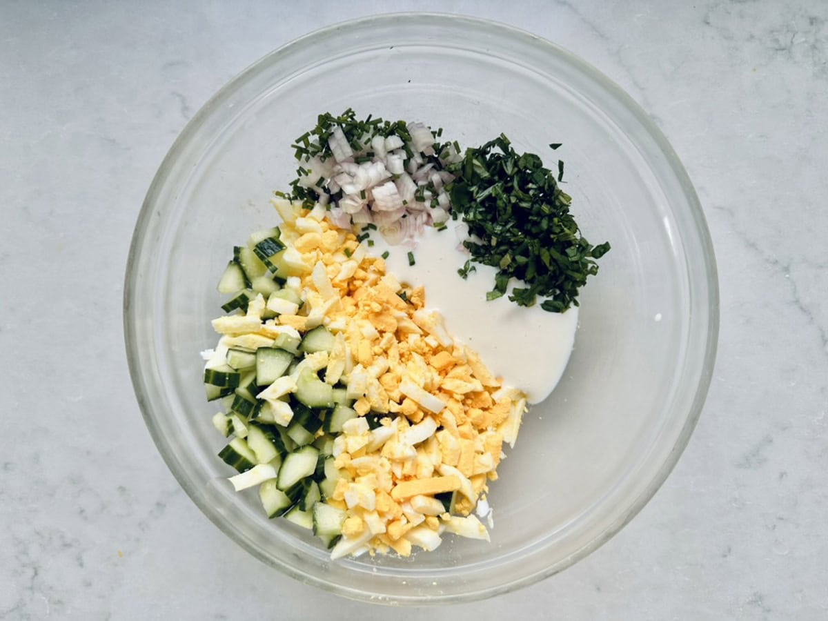 A glass bowl contains chopped cucumber, hard-boiled egg, red onion, and fresh basil with a white creamy dressing, all placed separately before mixing to create a vibrant Cucumber and Basil Egg Salad on a light countertop.