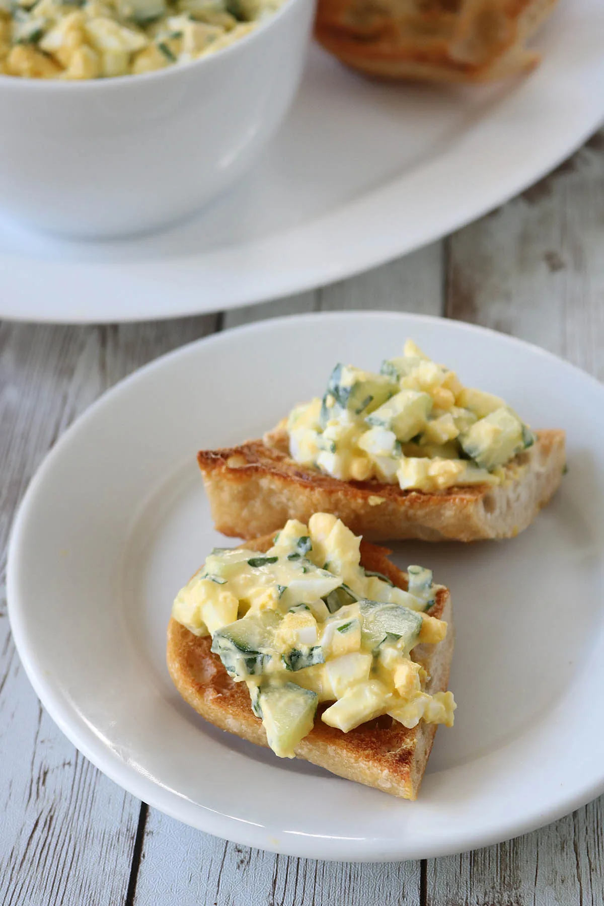 Two slices of toasted bread topped with Cucumber and Basil Egg Salad, placed on a white plate, with a bowl of egg salad in the background on a wooden surface.