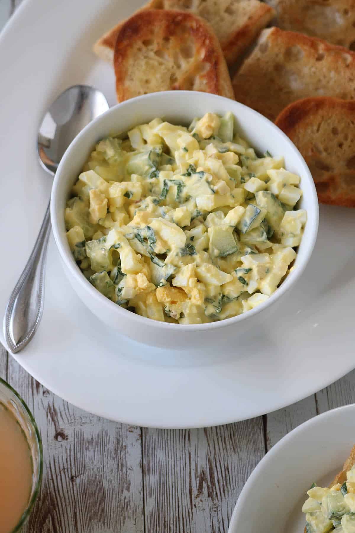 A bowl of Cucumber and Basil Egg Salad with herbs sits on a white plate next to toasted bread slices and a spoon on a rustic wooden table.