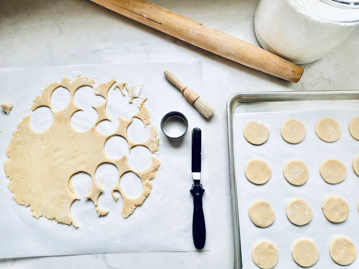 A baking scene with rolled cookie dough being cut into circles, a rolling pin, flour jar, brush, spatula, and a tray lined with parchment paper holding neatly arranged unbaked Strawberry Shortcake Cookies.