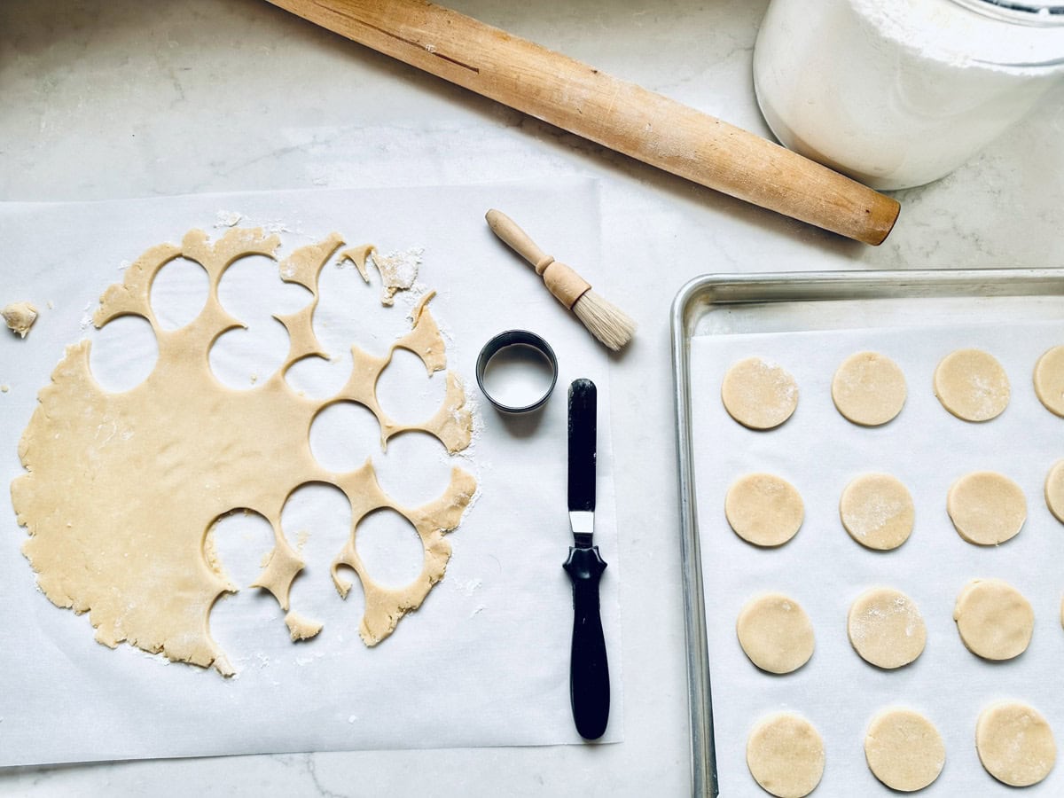 A baking scene with rolled cookie dough being cut into circles, a rolling pin, flour jar, brush, spatula, and a tray lined with parchment paper holding neatly arranged unbaked Strawberry Shortcake Cookies.