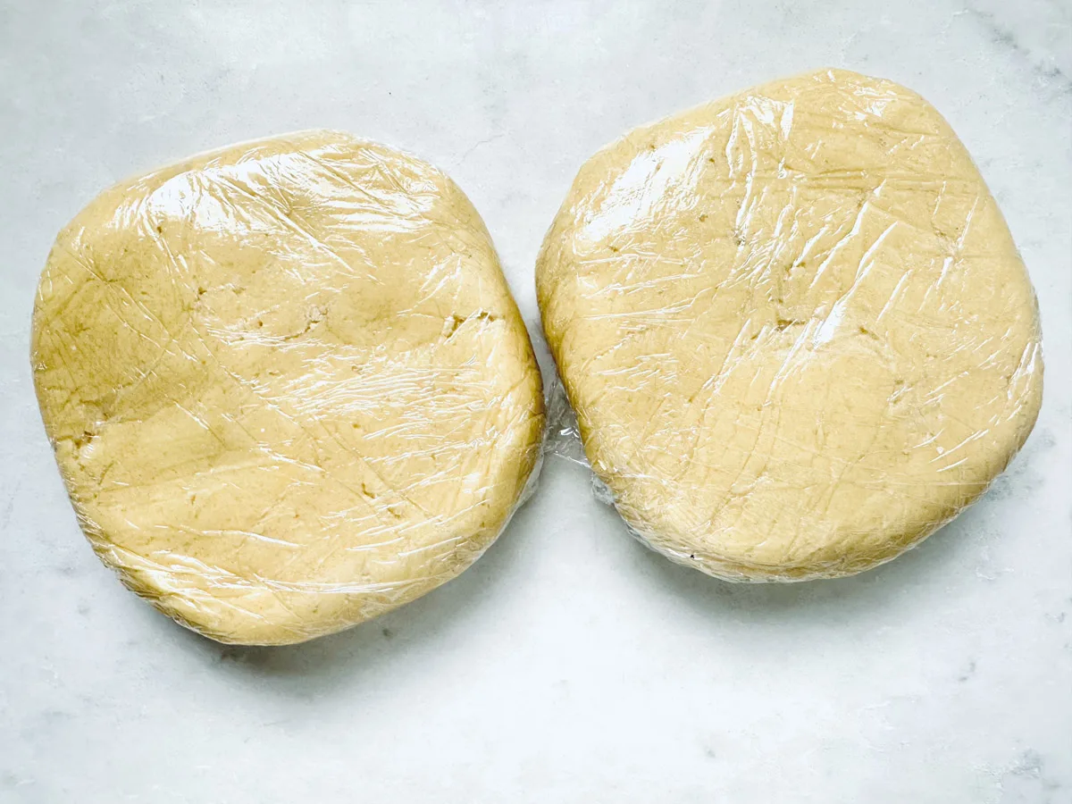 Two discs of dough for Strawberry Shortcake Cookies are wrapped in plastic wrap and placed side by side on a light-colored surface.