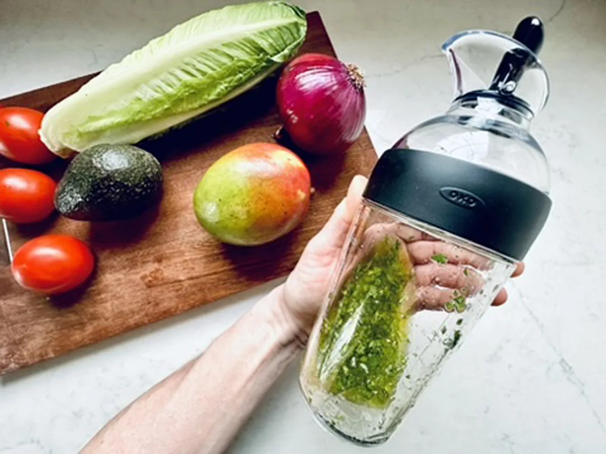 A hand holds a glass salad dressing mixer with green dressing inside, next to a cutting board piled with romaine lettuce, avocado, mango, red onion, and Roma tomatoes&mdash;perfect ingredients for a refreshing Mango, Tomato, and Avocado Salad.