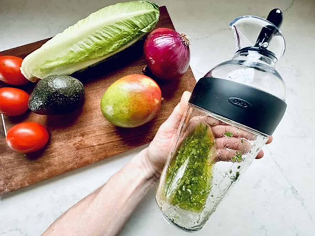 A hand holds a glass salad dressing mixer with green dressing inside, next to a cutting board piled with romaine lettuce, avocado, mango, red onion, and Roma tomatoes—perfect ingredients for a refreshing Mango, Tomato, and Avocado Salad.