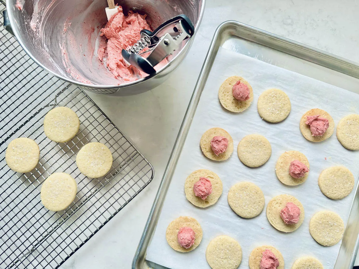 A baking scene with Strawberry Shortcake Cookies cooling on a rack, more cookies on a baking sheet topped with pink frosting, and a mixing bowl of pink frosting beside a cookie scoop and spatula.