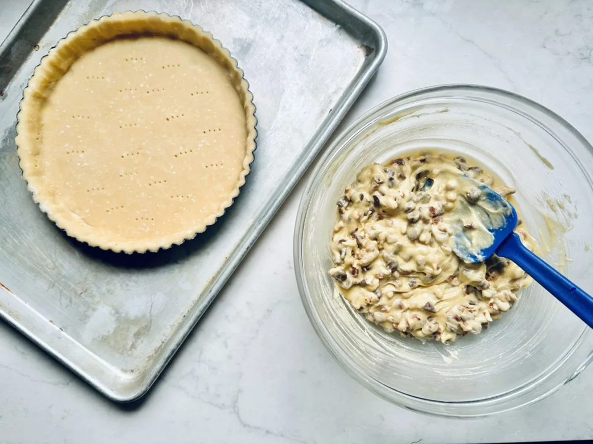 A tart crust with fork marks sits on a baking sheet next to a glass bowl of mixed filling, containing nuts and other ingredients, with a blue spatula resting inside.