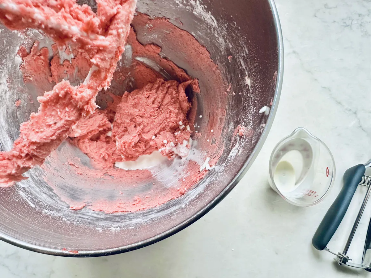 A mixing bowl with pink Strawberry Shortcake Cookies dough and a mixer paddle. Next to the bowl are a small glass measuring cup with milk and a cookie scoop on a light countertop.
