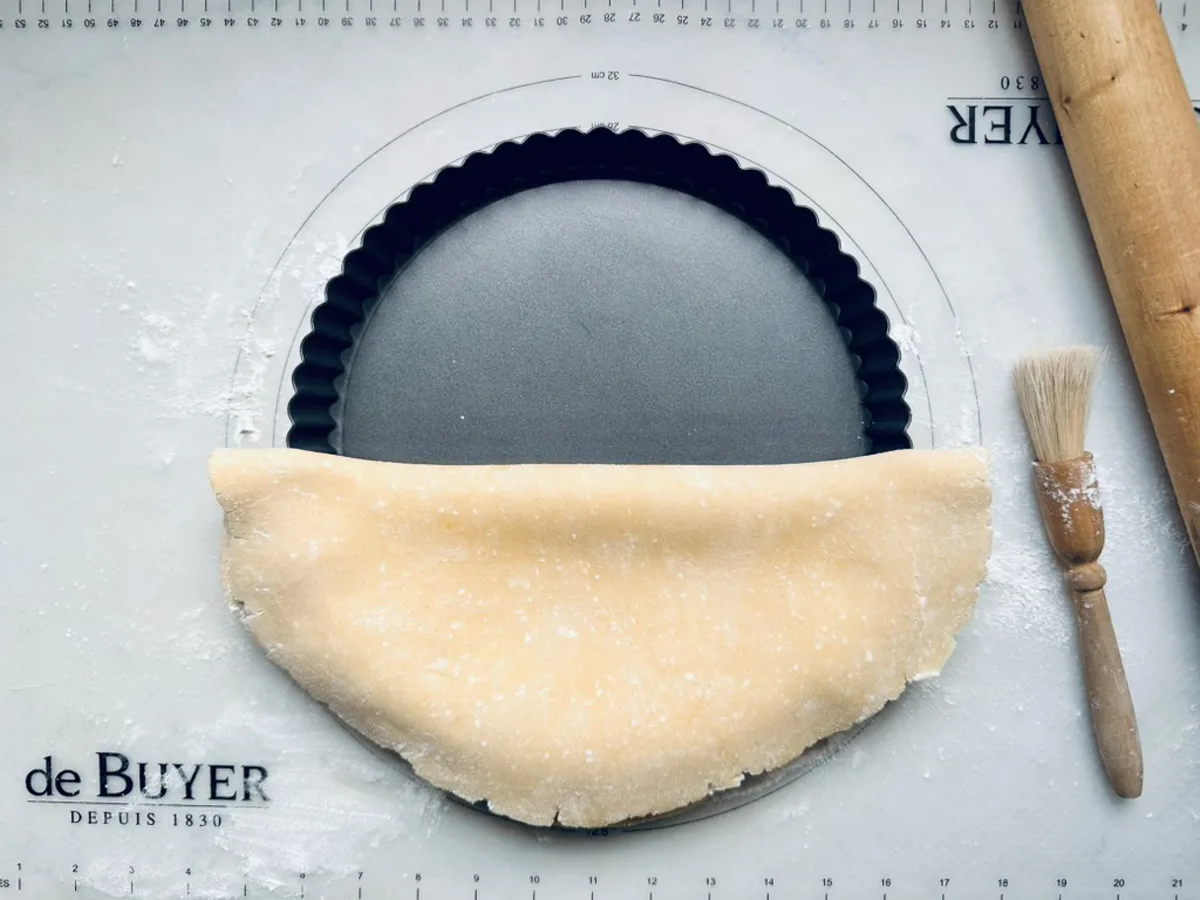A rolled-out sheet of dough is being placed over a round tart pan on a floured surface, with a rolling pin and pastry brush nearby.