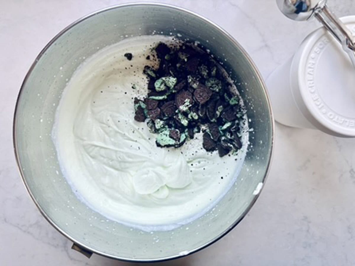 A mixing bowl containing light green whipped mixture with crushed chocolate mint cookies on top, next to an ice cream maker on a white surface.