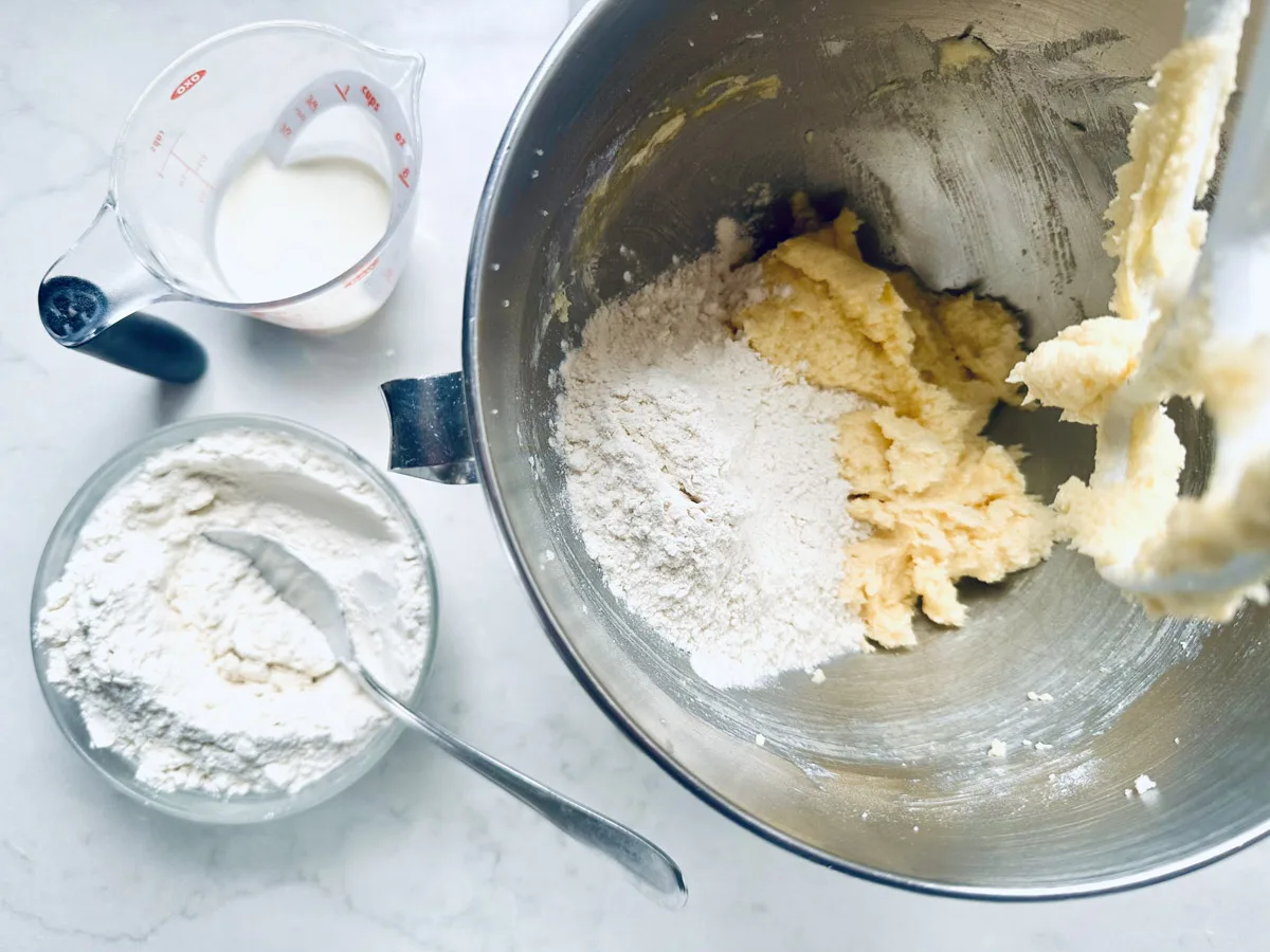 A mixing bowl with butter, sugar, and flour being creamed together for Strawberry Shortcake Cookies, next to a bowl of flour with a spoon and a measuring cup of milk on a white countertop.