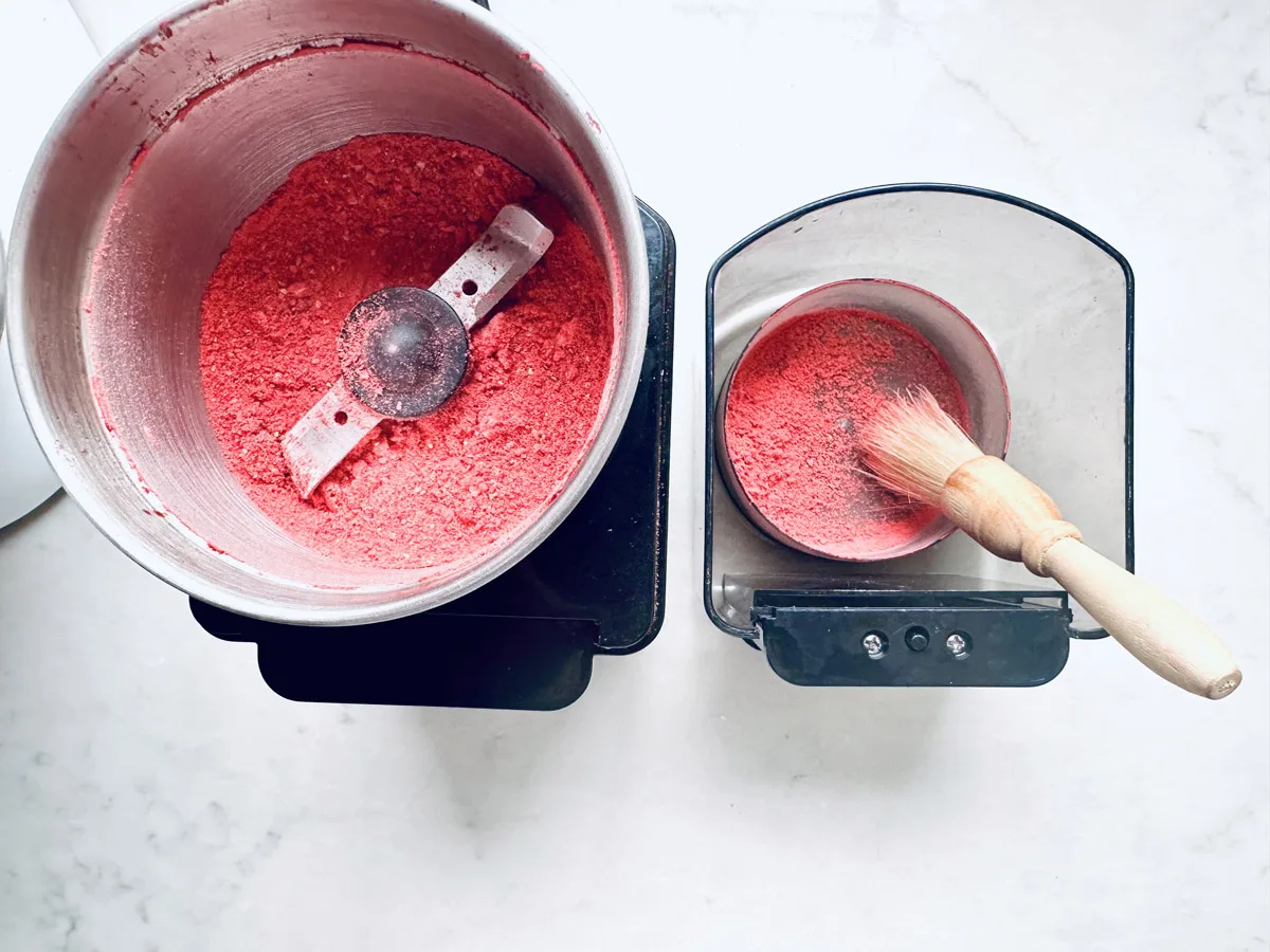 A metal grinder filled with bright red powder sits next to a small container holding some of the powder&mdash;perfect for dusting over Strawberry Shortcake Cookies&mdash;alongside a wooden-handled brush on a white surface.