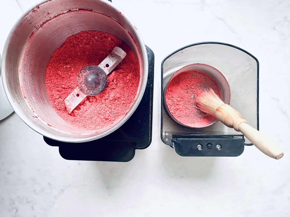 A metal grinder filled with bright red powder sits next to a small container holding some of the powder—perfect for dusting over Strawberry Shortcake Cookies—alongside a wooden-handled brush on a white surface.