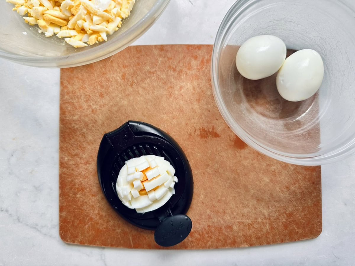 A brown cutting board with a sliced hard-boiled egg on an egg slicer, a clear bowl with two whole eggs, and a bowl of chopped eggs—everything you need to start making a fresh Cucumber and Basil Egg Salad—displayed on a white countertop.
