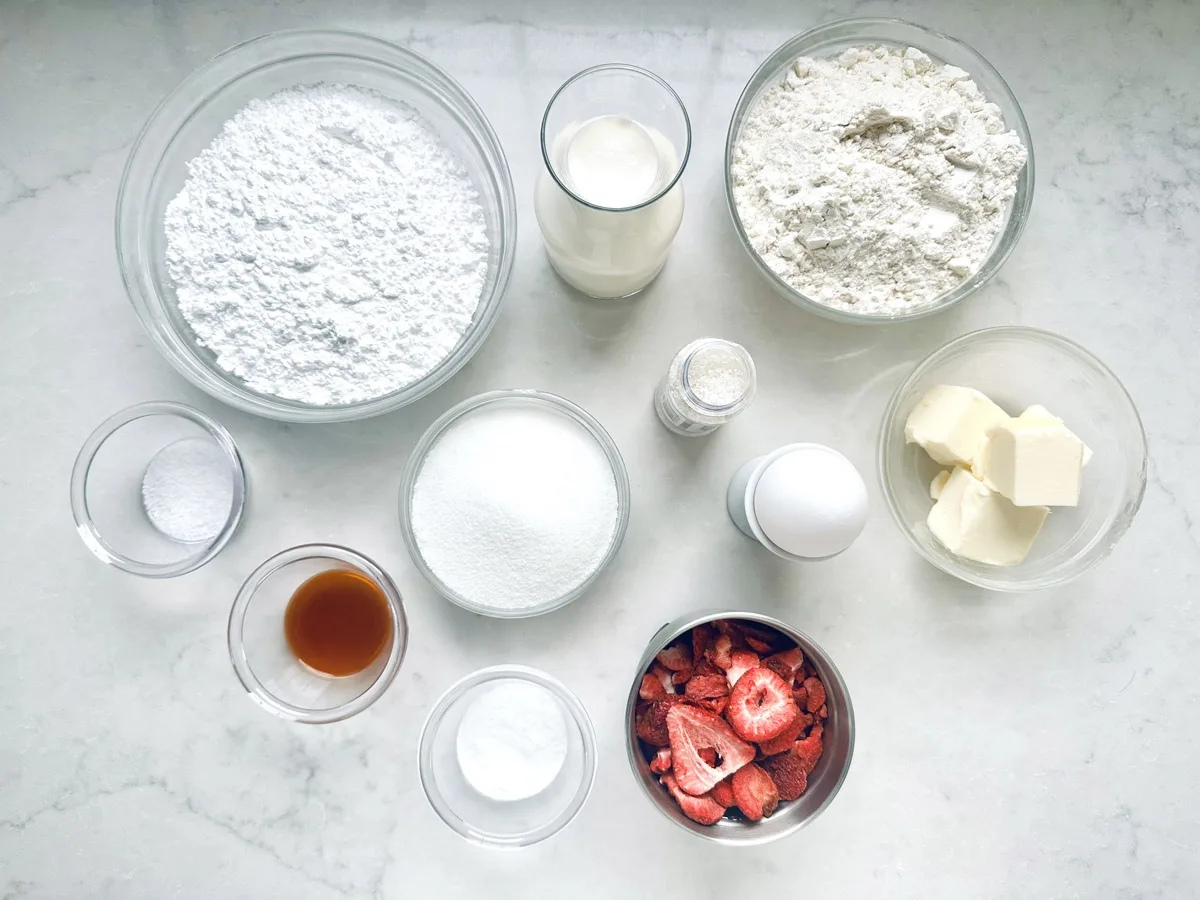A top-down view of baking ingredients for Strawberry Shortcake Cookies in bowls and glasses, including flour, powdered sugar, milk, butter, an egg, strawberries, vanilla, salt, baking powder, baking soda, and granulated sugar on a white surface.
