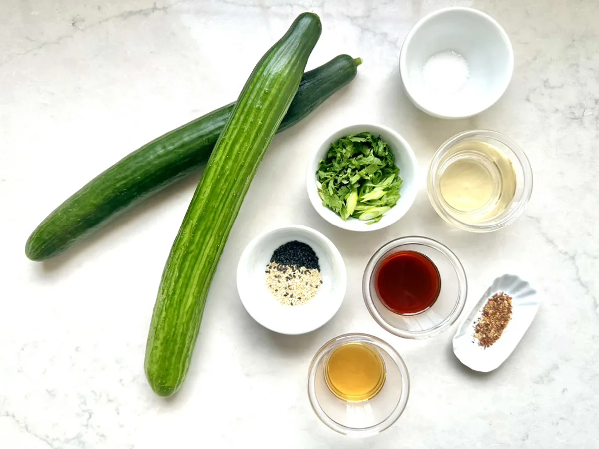 Two whole cucumbers and small bowls of salt, green onions, sesame seeds, rice vinegar, soy sauce, toasted sesame oil, and red pepper flakes are arranged on a white surface—perfect for making an Asian Cucumber Ribbon Salad.