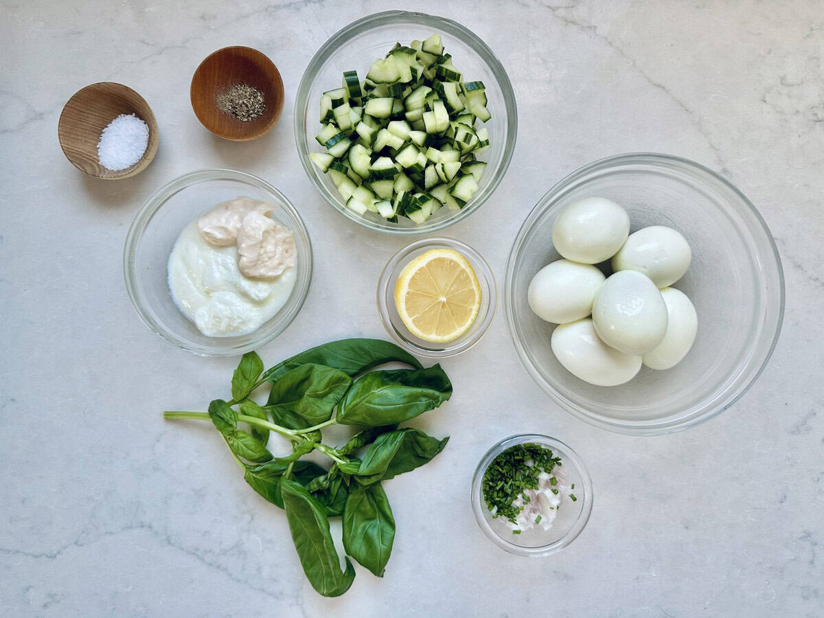 A marble countertop displays bowls of diced cucumber, hard-boiled eggs, mayonnaise and yogurt, chopped herbs with onions, salt and pepper, half a lemon, and fresh basil leaves—ingredients for a refreshing Cucumber and Basil Egg Salad arranged neatly.