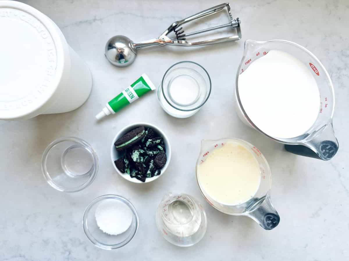 A flat lay of ingredients for mint cookies and cream ice cream: cream, sweetened condensed milk, glass of milk, sugar, Oreo cookies, mint extract, and a measuring cup, plus a metal ice cream scoop on a white surface.