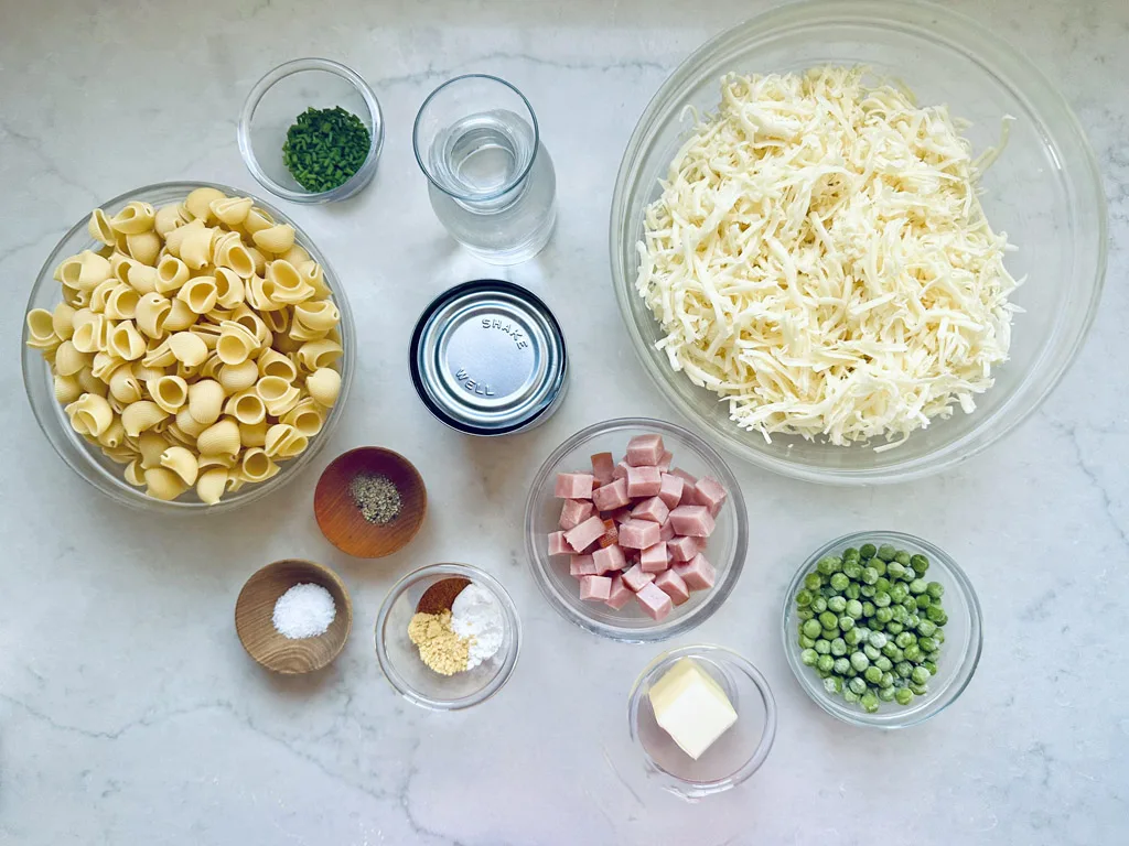 A top-down view of ingredients for a pasta dish, including dry pasta shells, shredded cheese, diced ham, green peas, butter, seasonings, herbs, and a glass of water, all arranged in glass bowls on a white surface.