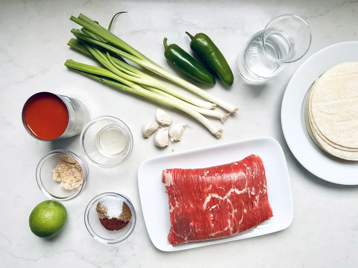 Top-down view of ingredients on a white surface: green onions, two jalape&ntilde;os, a glass of water, corn tortillas, a plate of raw steak, garlic cloves, a can of tomato sauce, lime, spices, and bowls of brown sugar and oil.