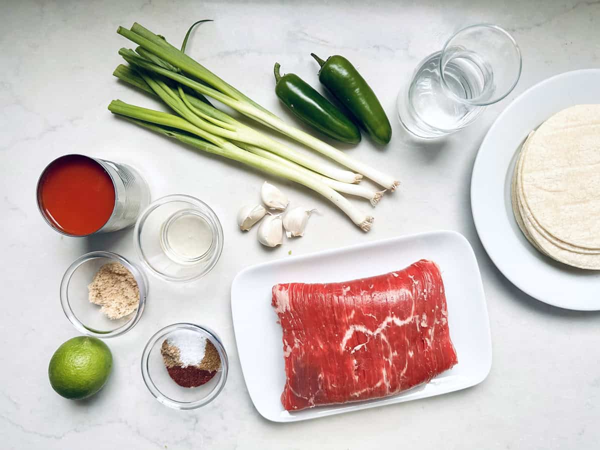 Top-down view of ingredients on a white surface: green onions, two jalapeños, a glass of water, corn tortillas, a plate of raw steak, garlic cloves, a can of tomato sauce, lime, spices, and bowls of brown sugar and oil.
