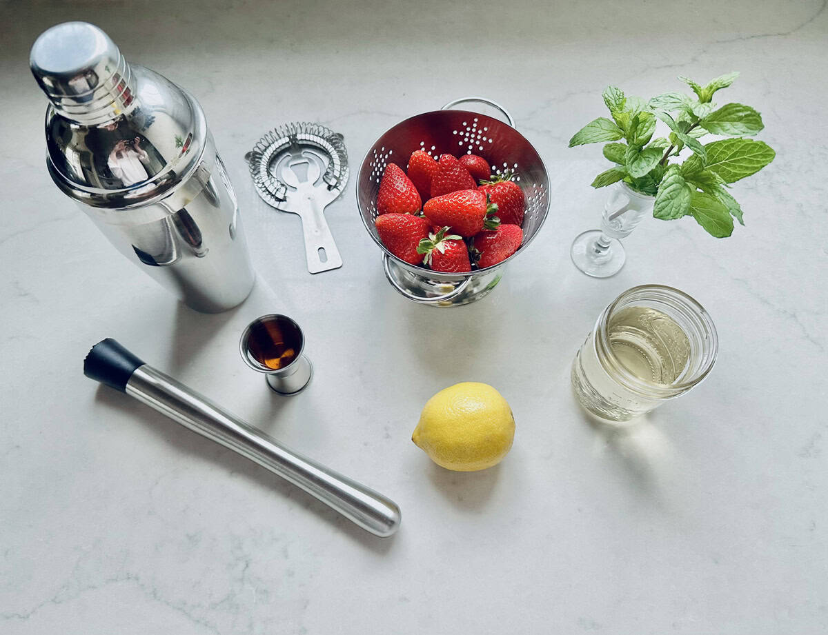 A cocktail shaker, strainer, fresh strawberries in a small colander, mint sprig in a glass, a jigger with dark liquid, muddler, lemon, and a jar with clear liquid arranged on a white countertop.