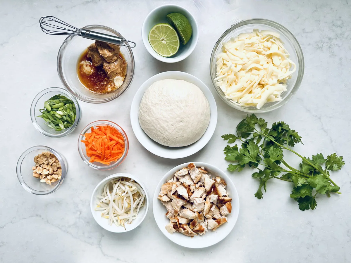 Assorted ingredients on a white surface, including dough, shredded cheese, cooked chicken pieces, sliced green onions, grated carrots, bean sprouts, chopped peanuts, fresh cilantro, a bowl of sauce with a whisk, and a halved lime.