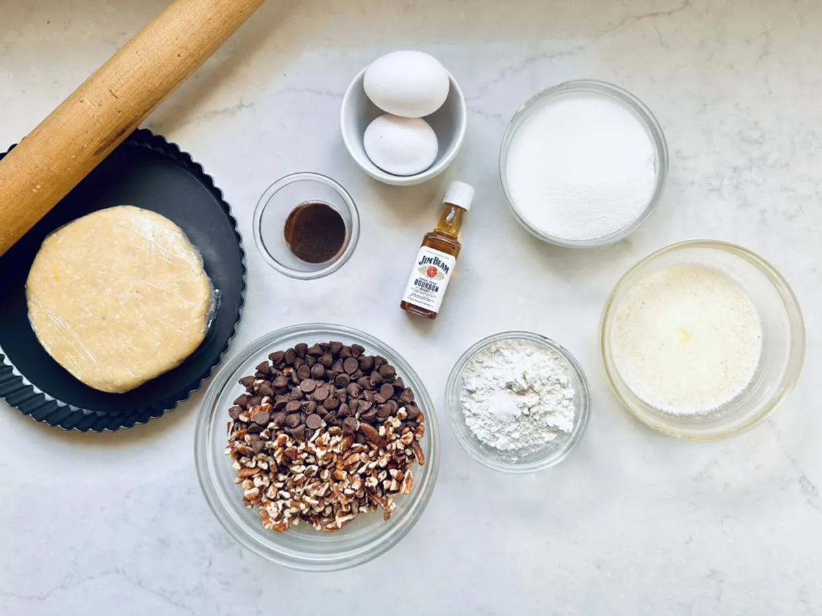 Baking ingredients on a marble surface: rolled pie dough, tart pan, two eggs, sugar, melted butter, chocolate chips, chopped pecans, flour, vanilla extract, and a small bottle of vanilla flavor. A rolling pin is nearby.